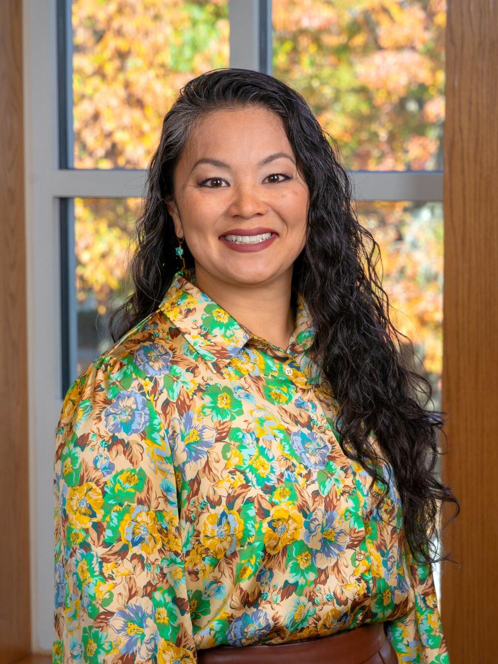 A woman smiles for a headshot wearing a colorful blouse in front of a bright window. She has long, dark, curly hair.
